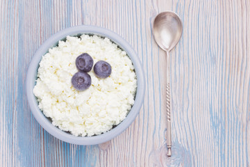 An overhead photo of fresh natural cottage cheese with blueberries and a silver spoon in a ceramic bowl on the wooden table. Organic eco healthy meal, dairy product. Top view with copyspase.