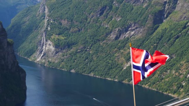 Norwegian flag waving on wind. Fjord Geirangerfjord in the background. View from Ornesvingen viewpoint, Norway