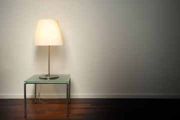 Modern design lamp on glass table illuminating minimalistic interior. Shaded wall with copy space and wooden floor.