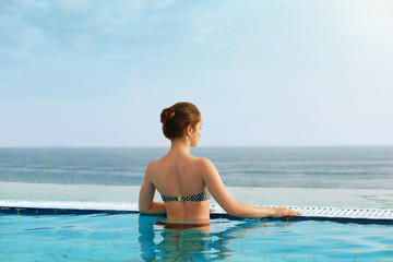 Fototapeta na wymiar Luxury Resort. Woman Relaxing In Infinity Swimming Pool Water. Beautiful Happy Healthy Female Model Enjoying Summer Travel Vacation, Looking At Sea View. Summertime Recreation, Relax And Spa Concept.