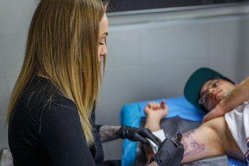 A boy with glasses and blue cap is tattooed by a young woman in her tattoo studio