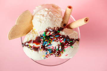 top view of vanilla flavor ice cream in glass bowl with chocolate sauce, strewed sprinkles and waffle cookies on pink background