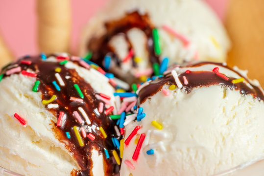close up of vanilla flavor ice cream in glass bowl with chocolate sauce, strewed sprinkles and cookies on pink background