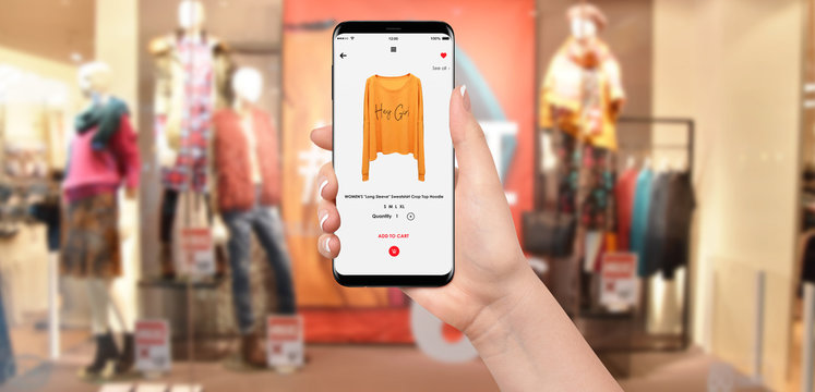 Woman In front of clothing store buying yellow hoodie online on her phone, blurred clothing store in background