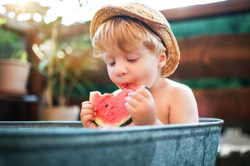Small boy with a hat in bath outdoors in garden in summer, eating watermelon.