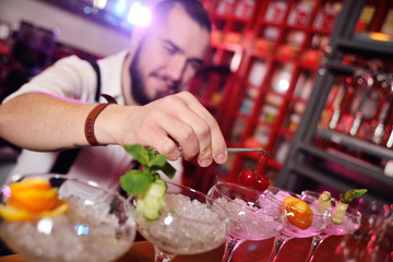 a handsome young bartender prepares cocktails and puts cherries for cocktails on the background of...
