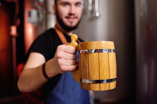 young bearded attractive male brewer with wooden beer mug in hand on background of brewery