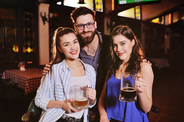 friends - young guy or man and two cute girls smiling holding glasses with beer on the background of the bar