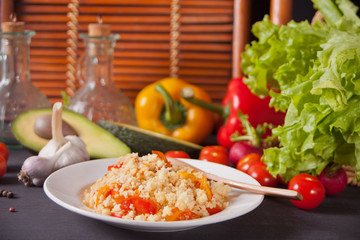 Delicious homemade vegetarian couscous with tomatoes, carrots, pepper and fresh basil on a dark kitchen table with vegetables on the background