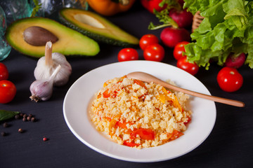 Delicious homemade vegetarian couscous with tomatoes, carrots, pepper and fresh basil on a dark kitchen table with vegetables on the background