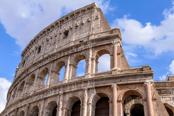 Fototapeta premium Wall of The Colosseum - A close-up low-angle view of upper section of west outer wall of the Colosseum, against white clouds and blue sky, on a sunny October afternoon. Rome, Italy.