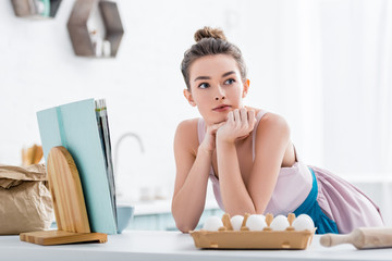 dreamy attractive girl near cookbook and eggs looking away