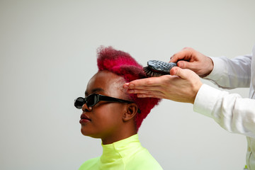 Picture showing african american woman at the hair salon. Studio shot of graceful young girl with stylish short haircut and colorful hair on gray background and hands of hairdresser.
