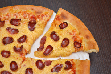 Pizza with cheese and smoked sausages on a white plate on a dark wooden table, one piece cut off, closeup