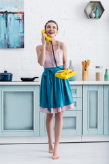 happy barefoot young woman talking on retro yellow telephone and laughing in kitchen