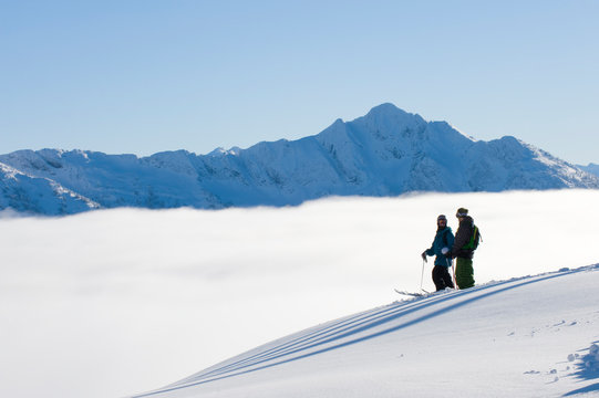 Mark Abma and Sean Pettit taking in the view while traversing on skiis above the clouds with mountain range in background in Mustang Powder Cats mountain area, BC, Canada.
