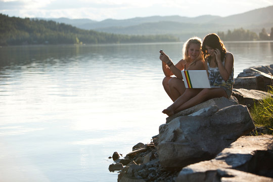 Two young women talk while texting and chatting on a computer on a sunny day at the lake.