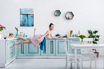 beautiful barefoot girl in elegant dress and apron levitating in air with pan in kitchen