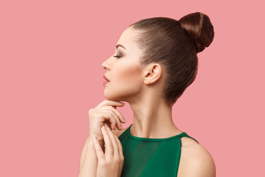 Profile side view portrait of calm serious beautiful young woman with bun hairstyle and makeup in green dress standing with closed eyes and touching her chin. studio shot, isolated on pink background.