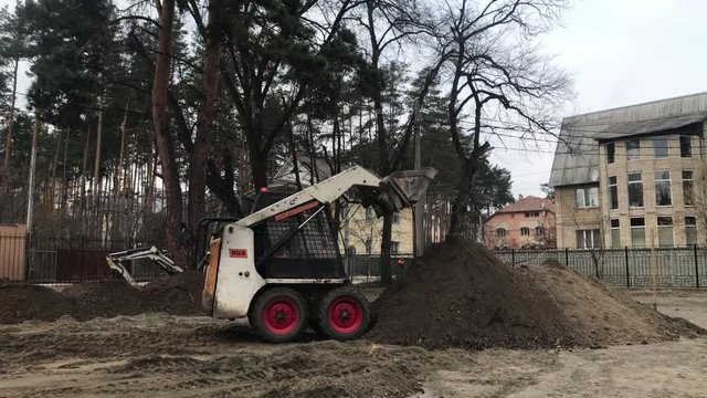 Two tractors in operation. Excavator digs the ground. Workers work on heavy machinery. Builders level the soil for laying paving slabs.