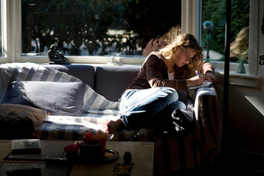 A young girl sitting on a sofa by a window writes in her journal.