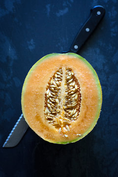 A halved Tuscan organic melon sits with a knife on a dark background.