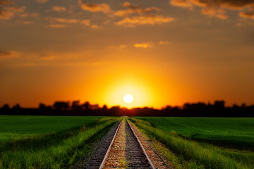 A railroad track dividing lush green agriculture fields under a bright orange and yellow sunset