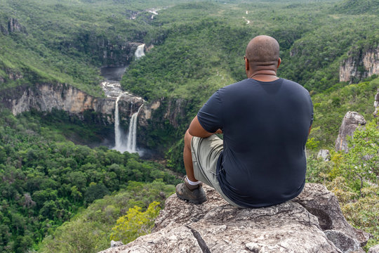 Mid adult hiker sitting on rocky edge looking at beautiful cerrado landscape with waterfalls and river in the green forest, Mirante da Janela peak, Chapada dos Veadeiros, Goias state, central Brazil