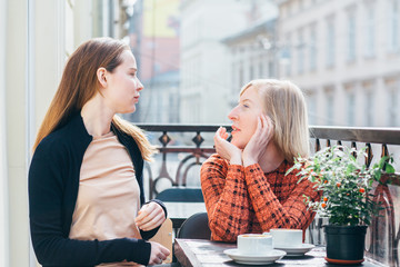 Two beautiful women, mature mother and adult daughter talking, relaxing on the balcony with city street view at spring sunny day. They drink coffee. Relationships and shared family time concept.