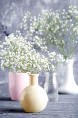 White gypsophila flowers in vases, cup and glass bottle on grey background