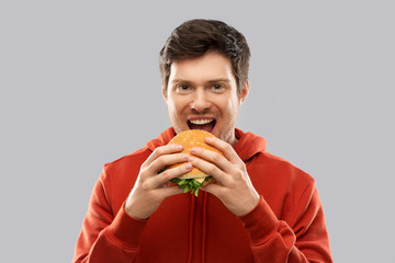 fast food and people concept - happy smiling young man in red hoodie eating hamburger over grey background