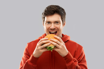 fast food and people concept - happy smiling young man in red hoodie eating hamburger over grey...