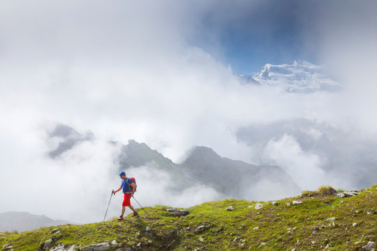 A male hiker on a grassy mountain ridge, with clouds around the Grand Combin.