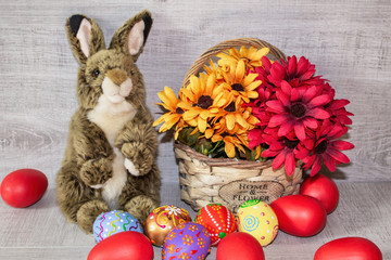 Colorful painted Easter eggs lie in a basket with flowers, on a wooden background, with a fluffy Easter bunny.