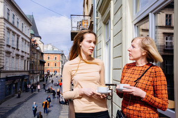 Two beautiful women, mature mother and adult daughter talking, standing on the balcony with city street view at spring sunny day. They drink coffee. Relationships and shared family time concept.