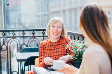 Happy smiling middle age blond mother and her adult daughter drinking coffee outdoor at sunny morning. Relationships and shared news family time concept.