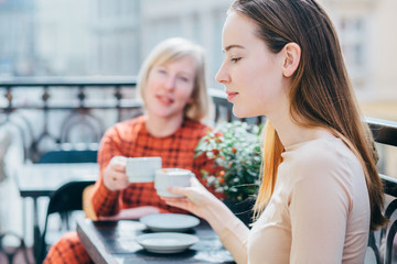 Obraz na płótnie Canvas Side view of charming adult daughter and her happy laughing middle age blond mother in orange dress on background drinking coffee outdoor at sunny morning. Relationships, family time concept.