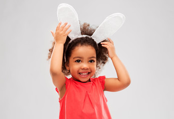 childhood, party props and easter concept - happy little african american girl wearing bunny ears headband over grey background