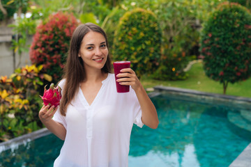 Portrait of happy girl with beautiful smile holds glass of dragon fruit smoothie and fruit in her hands background of pool