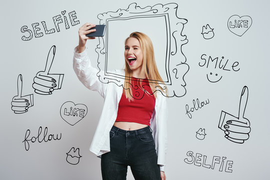 Selfie time! Cheerful young woman with blond hair making selfie by her smartphone and giving a wink while standing against grey background with different hand drawn doodle illustrations on it.