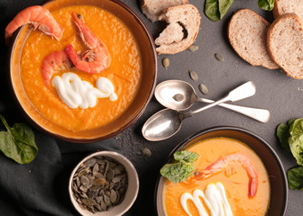 top view of orange pumpkin soup with shrimps, pumpkin seeds in dark bowls and bread, silver spoons on black background