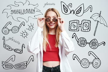 Beauty in sunglasses. Stylish and cheerful blonde woman in casual clothes wearing sunglasses while standing against grey background with hand drawn doodles on it.