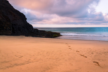 Foot prints in the sand on an empty beach leading towards the turquoise sea water under a beautiful sunrise sky. The Wax Beach (Playa de la Cera) tourist attraction in Lanzarote, Canary Islands. 
