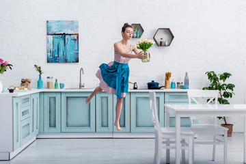 beautiful barefoot girl in elegant dress and apron flying in air with bouquet of tulips in glass...