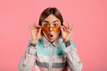 Amazed shocked young woman stand and look on camera. She hold sunglasses with hands. Isolated over...