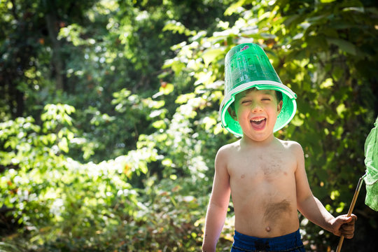 Toddler boy jokes with a bucket on his head and butterfly net in hand while next to creek in Bidwell Park, Chico, California.