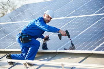 Man engineer in blue suit and helmet installing stand-alone solar photovoltaic panel system using screwdriver. Electrician mounting solar module on roof of modern house. Alternative energy concept.