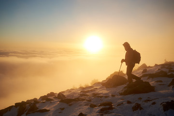 Silhouette of tourist hiker with backpack and trekking sticks hiking on rocky snowy mountain steep slope on background of foggy valley filled with white puffy clouds, raising sun and blue sky at dawn.