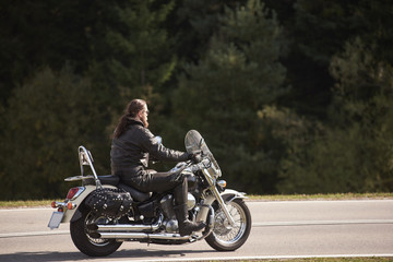 Fototapeta na wymiar Side view of bearded long-haired motorcyclist in sunglasses and black leather clothing riding motorbike along narrow asphalt path on sunny summer day on background of tall trees.