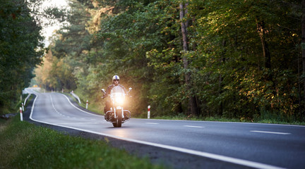 Handsome biker in white helmet riding motorcycle along asphalt road winding among tall green trees...